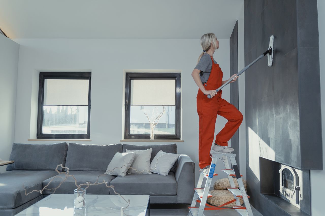 A woman in overalls cleans a wall in a stylish living room, improving the cleanliness of the home.