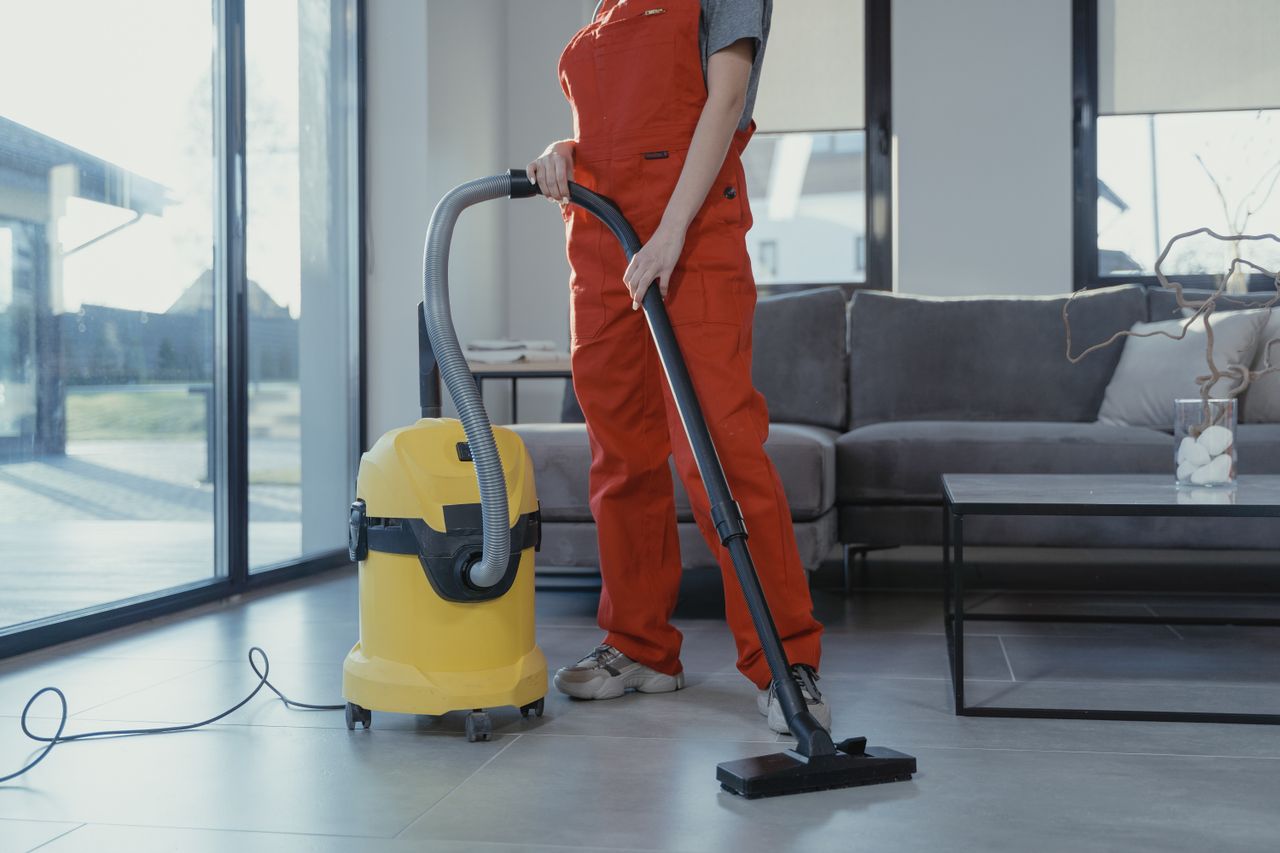 A professional cleaner in red coveralls vacuuming a contemporary living room with a yellow vacuum cleaner.